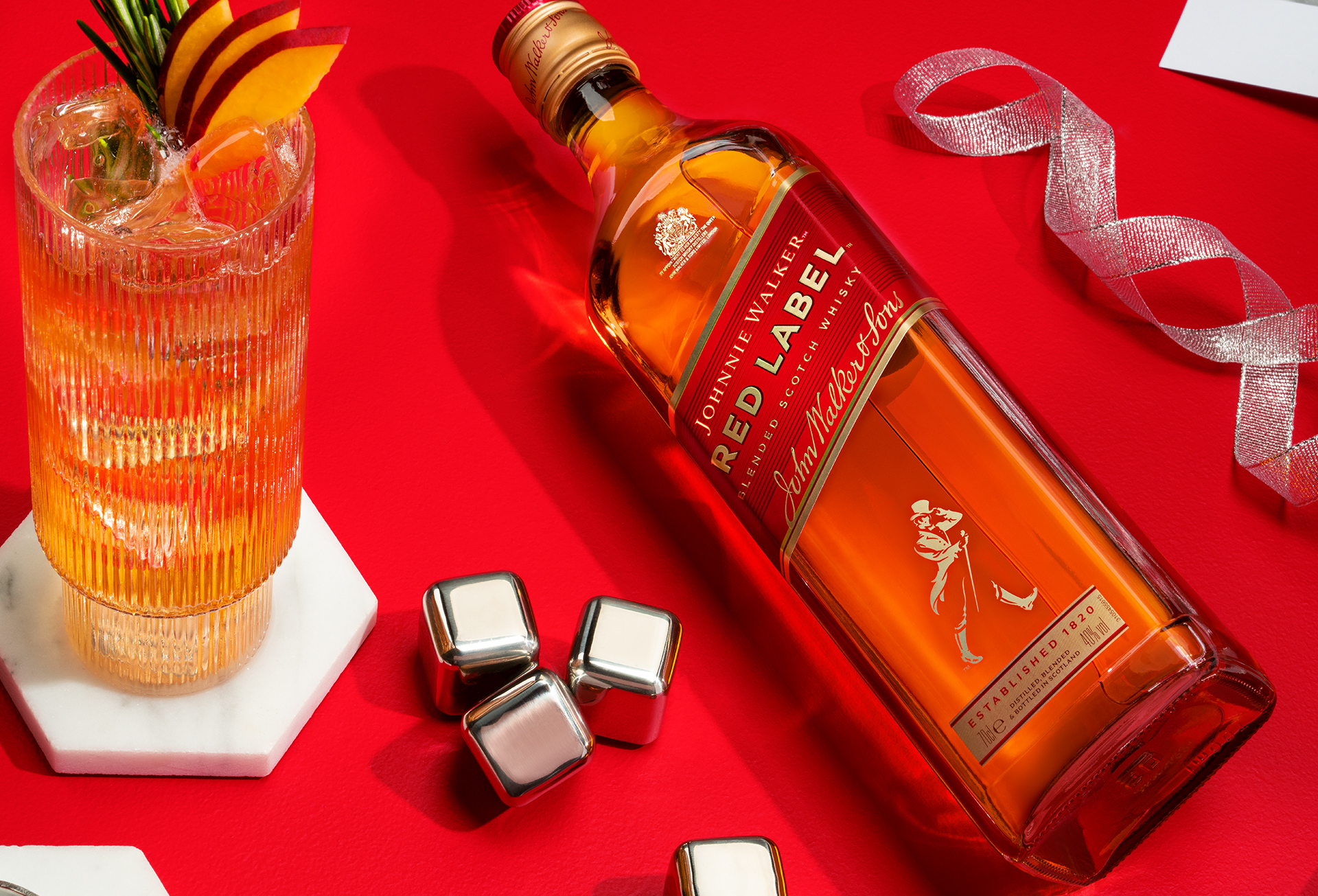 Garrafa de Johnnie Walker Red Label deitada sobre a mesa com fundo vermelho com um copo com gelo, whisky e pedaços de frutas ao lado, e ao redor fitas e pedras de gelo