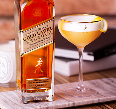 Garrafa do whisky Jonnie Walker Gold Label 1L a lado de uma taça com drink sobre a mesa