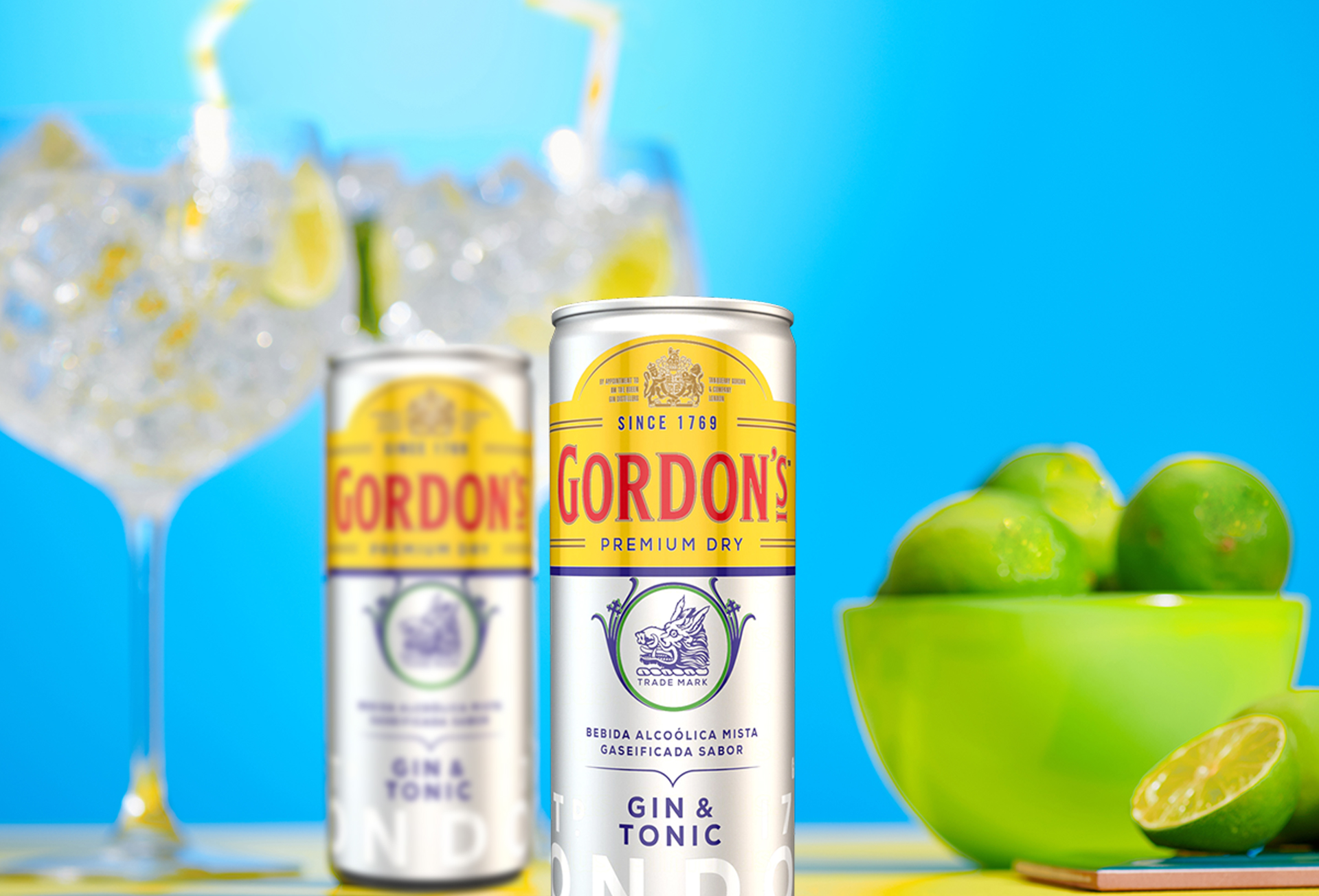 Duas latas de Gin Gordon`s london dry 269ml  em cima de uma mesa com duas taças com gelo e limões a mostra. 