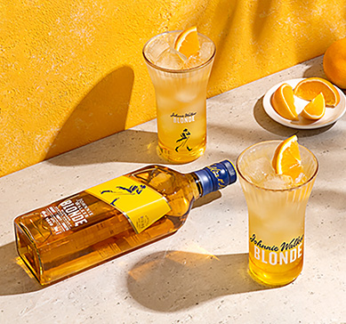 Garrafa do whisky JW Blonde sobre a mesa ao lado  de dois copos altos com laranja