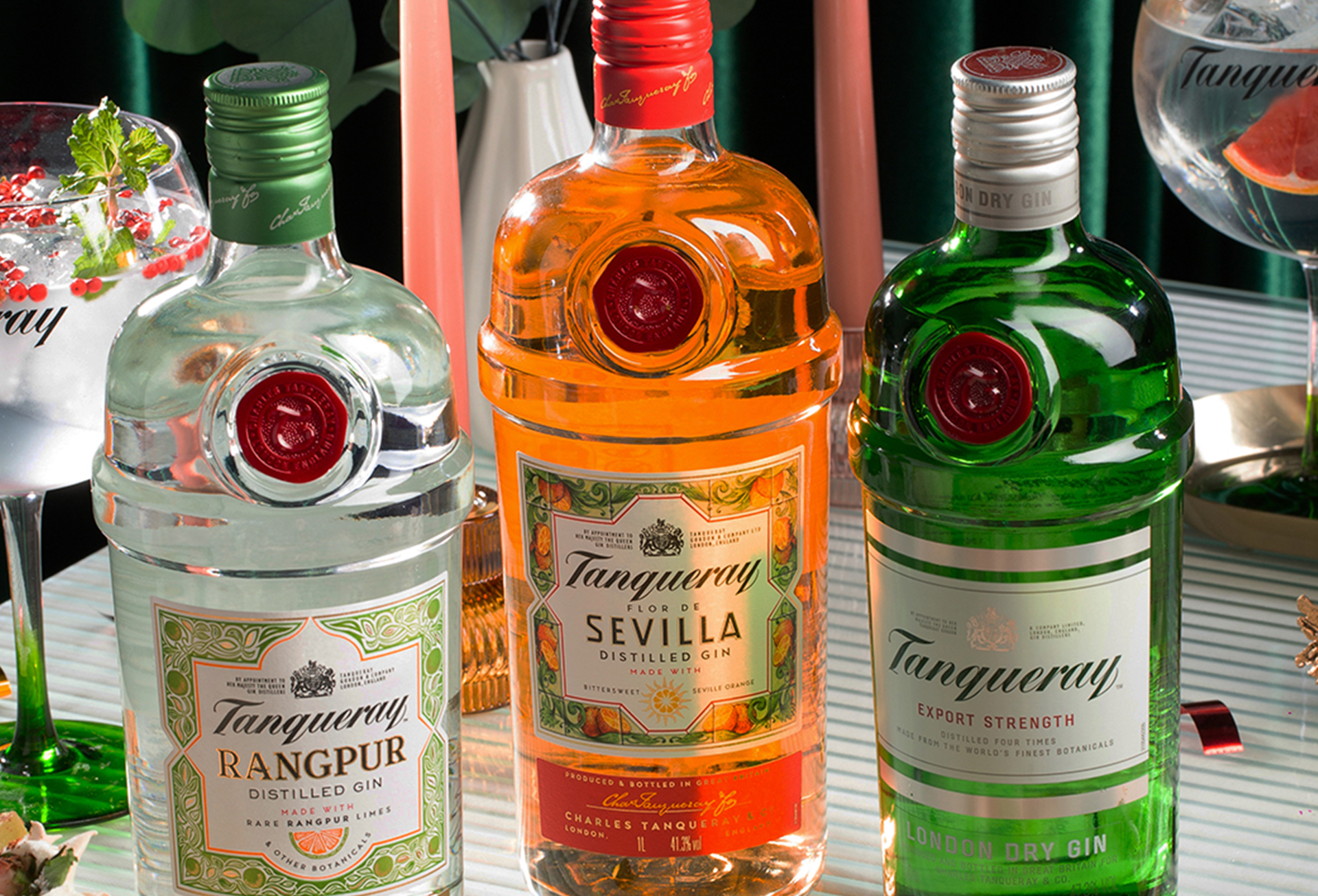 Três garrafas  Tanqueray estão lado a lado, com  drinks ao fundo, postos sobre uma mesa.