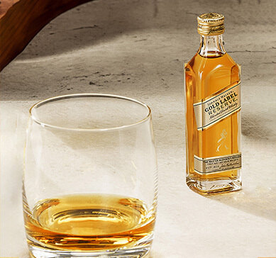 Mini garrafa de whisky Johnnie Walker Gold Label Reserve ao lado de copo com dose