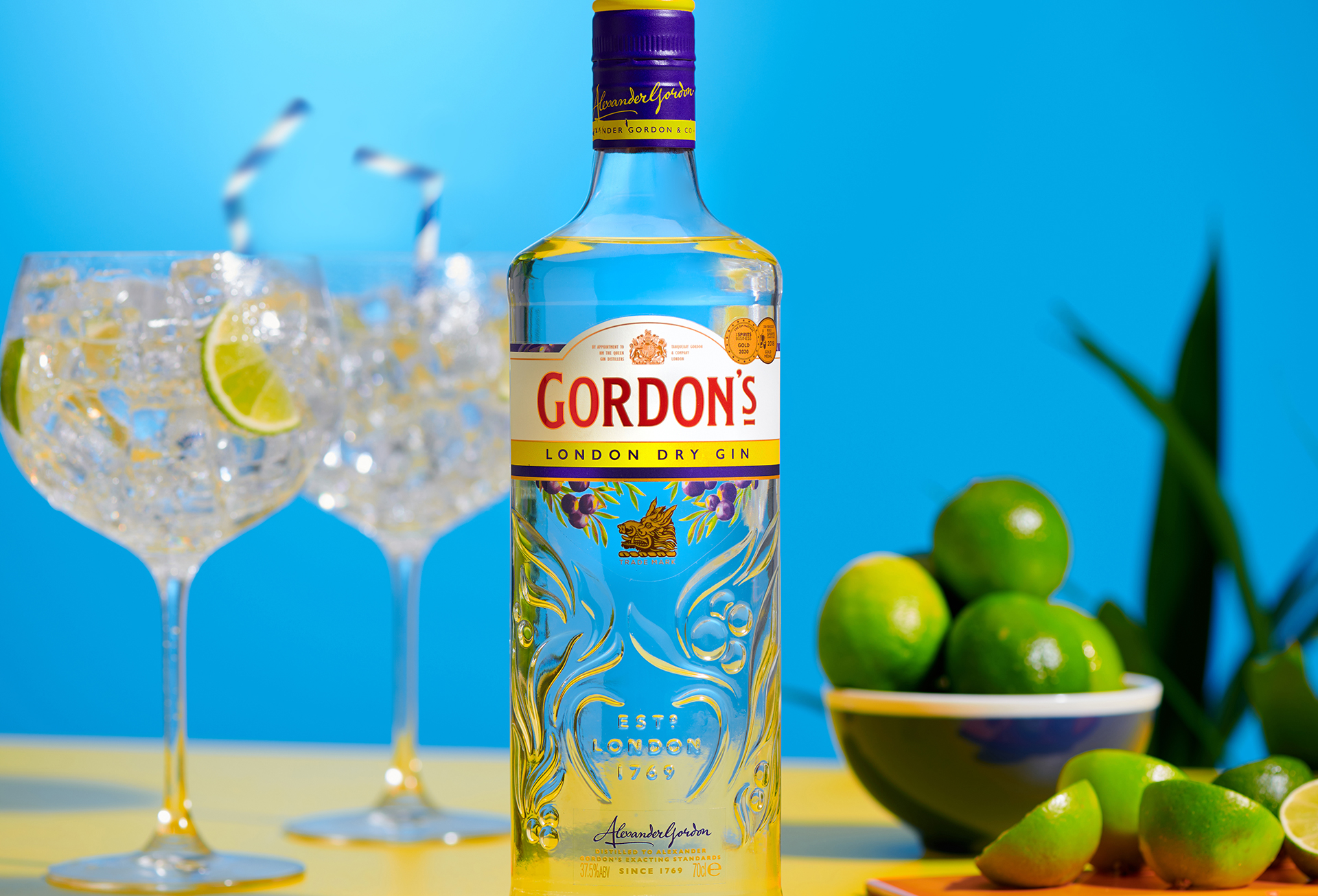 Garrafa Gordon's com duas taças ao fundo com gelo, gin, limão e canudo. Ao lado um bowl com limão